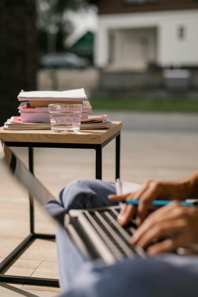 Close-up of hands typing on a laptop, glass of water, and notebooks outdoors, depicting remote education.